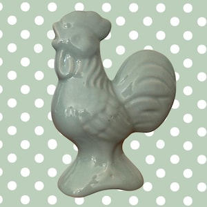 May include: A light blue ceramic rooster figurine. The rooster is standing upright with its beak open and a detailed comb. The figurine has a glossy finish and is set against a light green background with white polka dots.