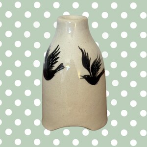 May include: A cream-colored ceramic vase with a wide base and a narrow neck. Two black bird silhouettes are painted on the front of the vase. The vase has a slightly irregular shape and a glossy finish.