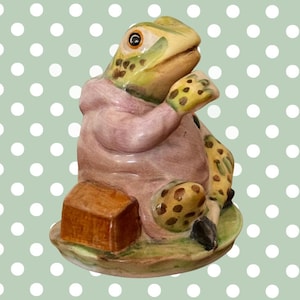 Könnte beinhalten: Eine Keramik-Froschfigur in einem rosafarbenen Kapuzenkleidungsstück, die auf einem runden Sockel neben einer kleinen braunen rechteckigen Schachtel sitzt. Der Frosch hat grüne Haut mit braunen Flecken, einen hellen Bauch und eine Hand am Gesicht.