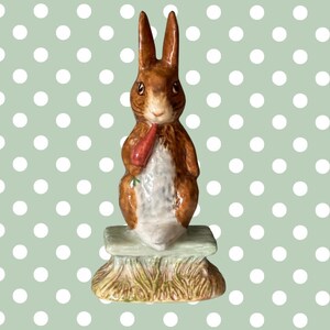 May include: A ceramic figurine of a brown rabbit holding a carrot, standing on a small, light green bench with a grassy base. The rabbit has long ears and a white belly. The background is a light green color with white polka dots.