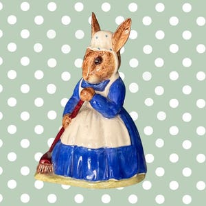 Pode incluir: Estatueta de cerâmica de um coelho com um vestido azul e um avental branco, segurando uma vassoura vermelha. O coelho tem pelo castanho e usa um chapéu branco. Fundo verde claro com bolinhas brancas.