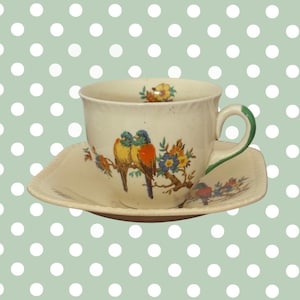 Könnte beinhalten: Cremefarbenes Tee-Set mit Vogel- und Blumendesign. Die Tasse hat einen grünen Henkel und die Untertasse ist quadratisch. Die Vögel sind gelb, orange und blau. Der Hintergrund ist hellgrün mit weißen Punkten.
