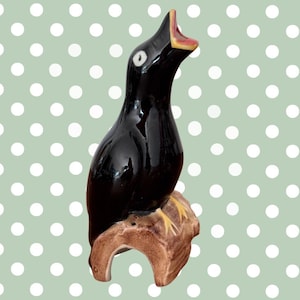 May include: A glossy black ceramic bird figurine with an open beak, perched on a brown base. The bird has a white eye and a red and yellow interior mouth. The base has a textured, bark-like appearance. The background is a light green with white polka dots.