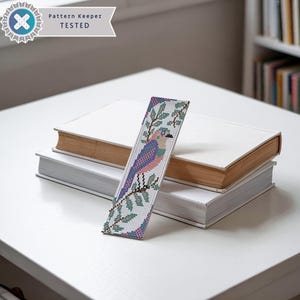 May include: A white beaded bookmark with a colorful bird and green leaves design. The bookmark is on top of two white books. The text "Pattern Keeper TESTED" is on a white label in the top left corner of the image.