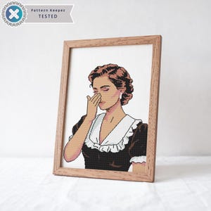 May include: A cross-stitch pattern of a woman with brown hair wearing a black and white dress, covering her nose and mouth with her hand. The pattern is framed in a light brown wood frame.