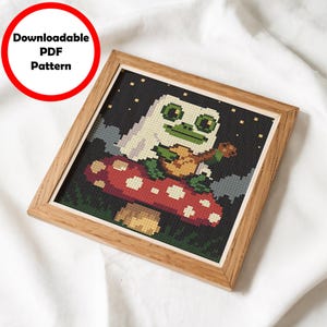 May include: Cross stitch pattern featuring a green frog wearing a white ghost costume, holding a guitar and sitting on a red and white mushroom. The background is a dark blue with white stars.