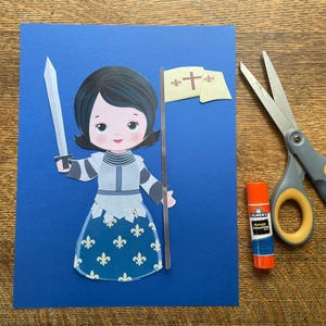 May include: A paper cutout of a cartoon knight holding a sword and a banner with a cross on a blue background. A pair of scissors and a glue stick are next to the cutout on a wooden surface.