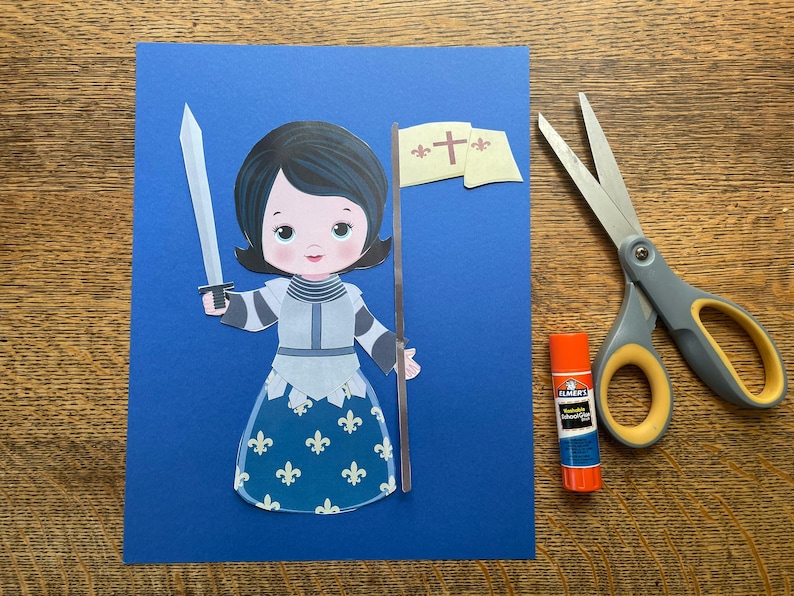 May include: A paper cutout of a cartoon knight holding a sword and a banner with a cross on a blue background. A pair of scissors and a glue stick are next to the cutout on a wooden surface.