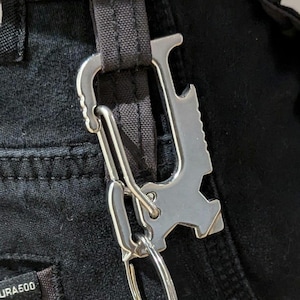 May include: A silver metal multi-tool keychain with a bottle opener, a carabiner clip, and a key ring. The keychain is attached to a black fabric strap.