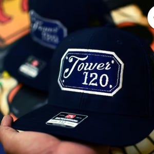 Puede incluir: Gorra de camionero azul marino con un parche con borde blanco que muestra el texto "Tower 120" en escritura blanca. La gorra tiene una parte trasera de malla y una visera curva. Otra gorra es visible en el fondo.