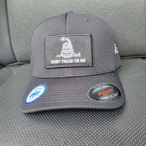 Peut inclure: Casquette de baseball noire avec une visière incurvée. Elle présente un écusson rectangulaire avec un serpent enroulé blanc et l'inscription "DON'T TREAD ON ME". La casquette a également une étiquette Flexfit.