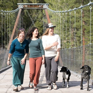 May include: Three women and two dogs walking across a suspension bridge. The women are wearing casual clothing and smiling. The dogs are black and white and brown and white.