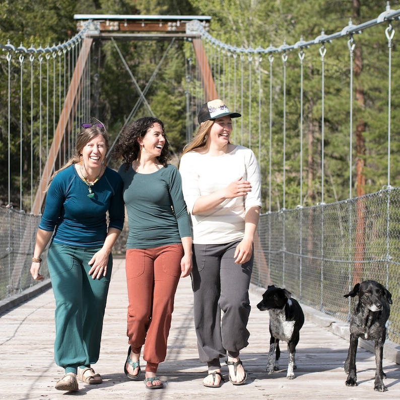 May include: Three women and two dogs walking across a suspension bridge. The women are wearing casual clothing and smiling. The dogs are black and white and brown and white.