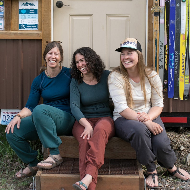 May include: Three women are sitting on a wooden porch, smiling and looking at the camera. They are wearing casual clothing, including a blue shirt, a green shirt, and a white shirt with a striped pattern. The woman in the middle is wearing a hat with a colorful design. The porch is in front of a wooden building with a door and a shelf with skis.