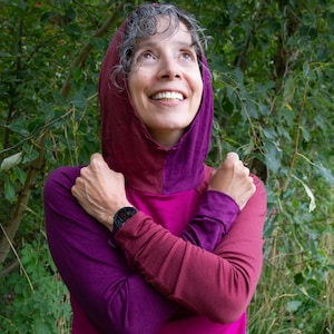 May include: A long-sleeved hooded shirt featuring a color-blocked design in shades of magenta, deep purple, and burgundy. The garment includes a fitted hood and long sleeves with a solid, lightweight fabric construction, worn with arms crossed.