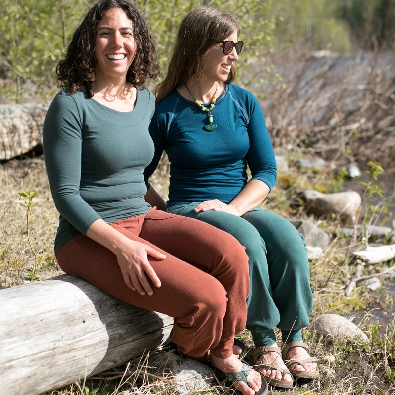 May include: Two women are sitting on a log in a forest setting. The woman on the left is wearing a teal long-sleeved shirt and rust-colored pants. The woman on the right is wearing a teal long-sleeved shirt and dark green pants. Both women are wearing sandals.