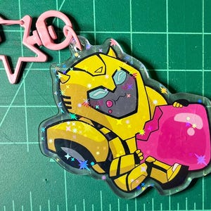 May include: A clear acrylic charm featuring a yellow and black cartoon robot character holding a pink object. The charm has a pink star-shaped clasp and a pink clip. The charm is decorated with glitter.