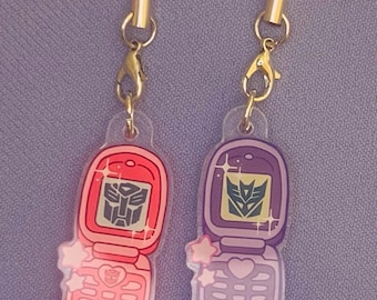 May include: Two clear acrylic cell phone charms, one red and one purple, each with a gold clasp and a red or purple cord. The charms feature a Transformers logo and star accents. The phone keypads have heart and circle designs.