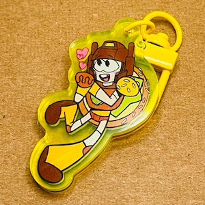 May include: A yellow acrylic keychain featuring a cartoon character in a burger costume. The character has a brown helmet, white face, and is surrounded by a yellow border. The keychain includes a yellow clasp and a small loop for attachment.