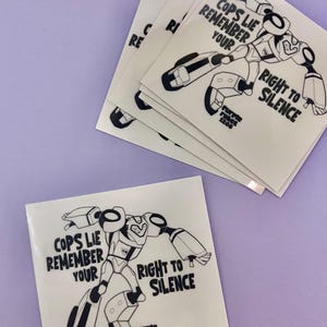May include: Black and white sticker with a robot illustration and the text "Cops Lie Remember Your Right to Silence". The sticker also includes the text "Purple Robot 1208".