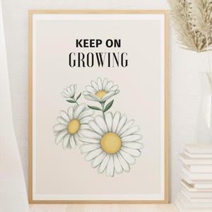 May include: A framed print featuring the words "KEEP ON GROWING" in black text above a watercolor illustration of daisies. The daisies have white petals and yellow centers. The frame is a light wood color.