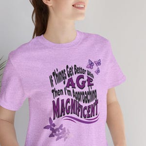 Könnte beinhalten: Ein helles lila T-Shirt mit dem Text "If things get better with age then I'm approaching magnificent" in Schwarz und Lila. Der Text ist von lila Schmetterlingen umgeben.