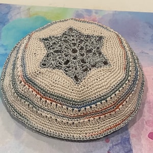 Peut inclure: Une kippa crochetée de couleur crème avec une bordure métallique argentée et un motif d'étoile de David au centre. La kippa présente des rayures bleues, orange et argentées. Cette coiffe religieuse est un article juif traditionnel.