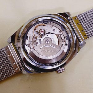 K&ouml;nnte beinhalten: Nahaufnahme der R&uuml;ckseite einer silberfarbenen Armbanduhr mit Mesh-Armband. Das Innenleben der Uhr ist sichtbar, einschlie&szlig;lich Zahnr&auml;dern, einem Rotor und dem Markennamen "SEIKO". Die Uhr liegt auf einer hellbeigen Oberfl&auml;che.