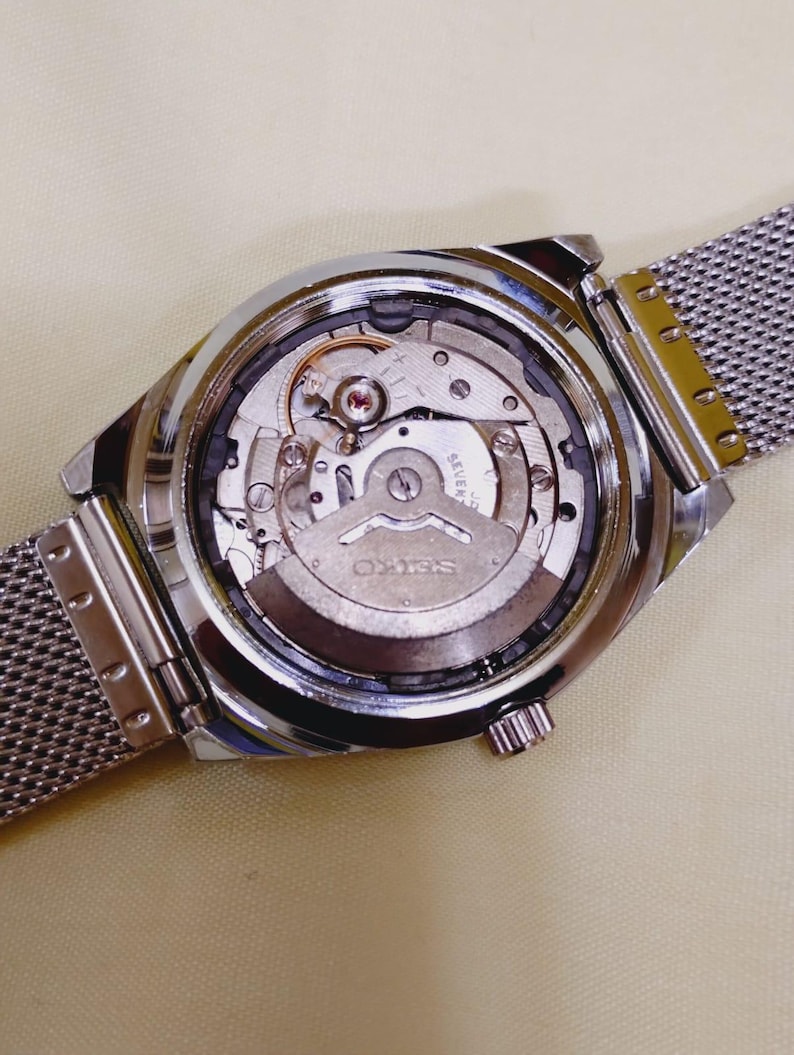 K&ouml;nnte beinhalten: Nahaufnahme der R&uuml;ckseite einer silberfarbenen Armbanduhr mit Mesh-Armband. Das Innenleben der Uhr ist sichtbar, einschlie&szlig;lich Zahnr&auml;dern, einem Rotor und dem Markennamen "SEIKO". Die Uhr liegt auf einer hellbeigen Oberfl&auml;che.