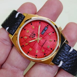Könnte beinhalten: Goldfarbene Seiko 5 Automatik-Uhr mit rotem Zifferblatt und schwarzem Lederarmband. Das Zifferblatt zeigt "TUE 13". Das Armband ist schwarz mit weißen Nähten. Eine klassische Uhr.