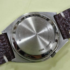 May include: Close-up of a silver-toned watch back with a brown leather strap. The watch back has etched text including "SEIKO", "WATER RESISTANT", and other details. The strap has white stitching.