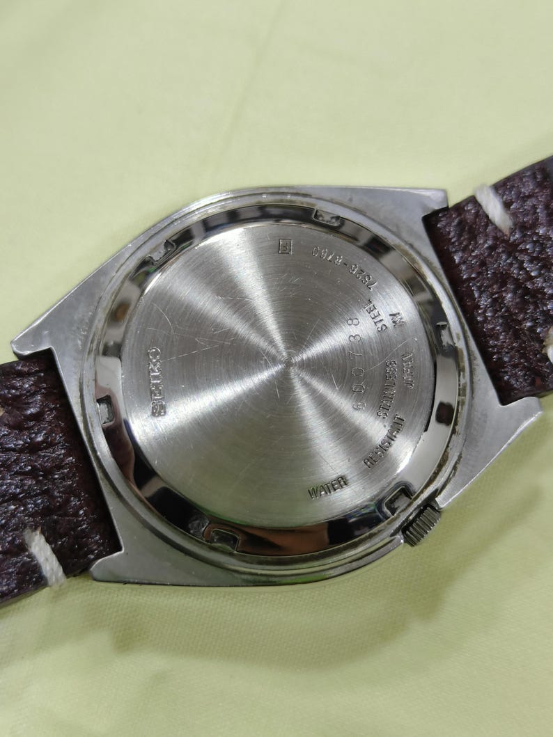 May include: Close-up of a silver-toned watch back with a brown leather strap. The watch back has etched text including "SEIKO", "WATER RESISTANT", and other details. The strap has white stitching.