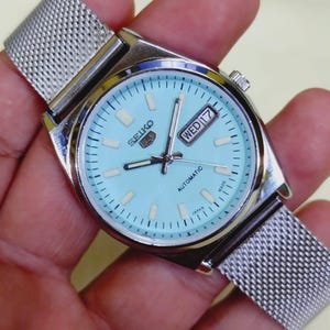 K&ouml;nnte beinhalten: Eine silberfarbene Seiko Automatik-Armbanduhr mit hellblauem Zifferblatt und Mesh-Armband. Das Zifferblatt zeigt Tag und Datum, mit dem Wort "Automatic" darunter. Die Uhr wird in Japan hergestellt.