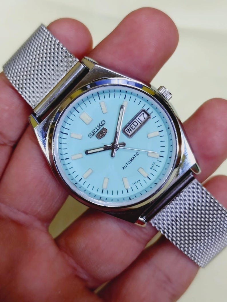 K&ouml;nnte beinhalten: Eine silberfarbene Seiko Automatik-Armbanduhr mit hellblauem Zifferblatt und Mesh-Armband. Das Zifferblatt zeigt Tag und Datum, mit dem Wort "Automatic" darunter. Die Uhr wird in Japan hergestellt.