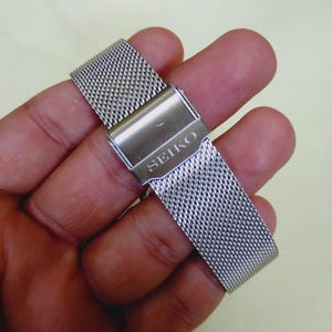 K&ouml;nnte beinhalten: Ein silberfarbenes Edelstahl-Uhrenarmband mit einem Mesh-Design. Das Armband hat einen Verschluss mit dem eingravierten Markennamen "SEIKO". Das Armband wird in einer Hand gehalten.