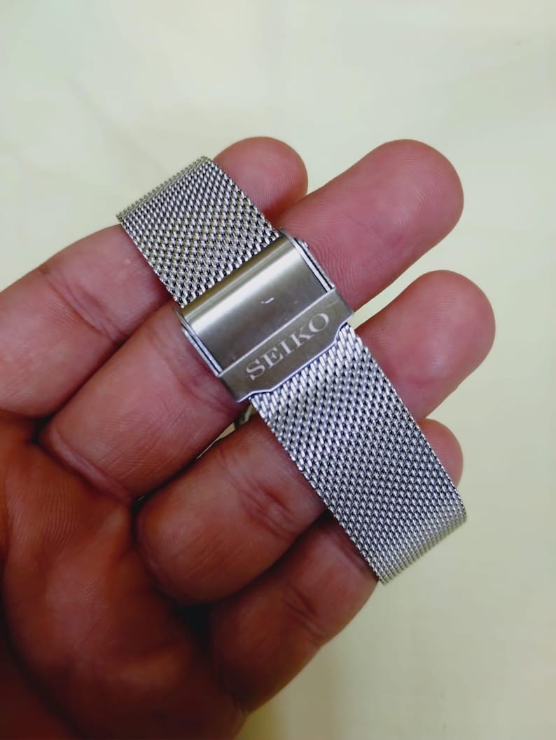 K&ouml;nnte beinhalten: Ein silberfarbenes Edelstahl-Uhrenarmband mit einem Mesh-Design. Das Armband hat einen Verschluss mit dem eingravierten Markennamen "SEIKO". Das Armband wird in einer Hand gehalten.