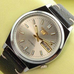 Op de afbeelding: Een zilveren Seiko automatische polshorloge met een zwarte leren band. De wijzerplaat van het horloge is zilver met zwarte markeringen en een goudkleurig dag- en datumavenster dat "WED10" aangeeft.