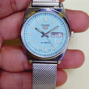 K&ouml;nnte beinhalten: Eine silberfarbene Seiko 5 Automatik-Uhr mit hellblauem Zifferblatt und Mesh-Armband. Das Zifferblatt zeigt Tag und Datum mit dem Text "SEIKO 5 AUTOMATIC". Die Uhr ist mit "JAPAN MADE" gekennzeichnet.
