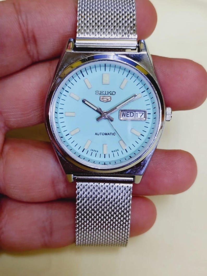K&ouml;nnte beinhalten: Eine silberfarbene Seiko 5 Automatik-Uhr mit hellblauem Zifferblatt und Mesh-Armband. Das Zifferblatt zeigt Tag und Datum mit dem Text "SEIKO 5 AUTOMATIC". Die Uhr ist mit "JAPAN MADE" gekennzeichnet.