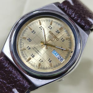 May include: A vintage Seiko automatic watch with a gold-toned face and a brown leather strap. The watch face displays the day as 'SAT 26' and features a grid pattern with hour markers. The watch case is silver-toned.