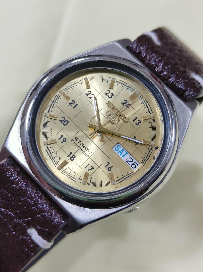 May include: A vintage Seiko automatic watch with a gold-toned face and a brown leather strap. The watch face displays the day as 'SAT 26' and features a grid pattern with hour markers. The watch case is silver-toned.