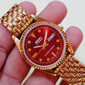 May include: A gold-coloured Seiko automatic watch with a red dial and a gold bracelet. The watch face displays the day as "SAT 28" and the text "Diver's 200m". The watch has gold hour markers and hands.