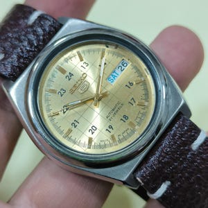 May include: A vintage Seiko 5 automatic watch with a gold-tone dial and a brown leather strap. The watch face displays the day and date, with the day reading "SAT 26". The watch has a silver-tone case and a grid pattern on the dial.