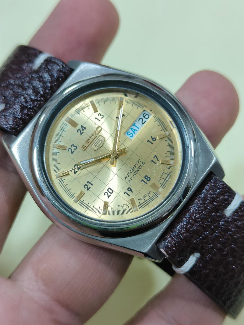 May include: A vintage Seiko 5 automatic watch with a gold-tone dial and a brown leather strap. The watch face displays the day and date, with the day reading "SAT 26". The watch has a silver-tone case and a grid pattern on the dial.