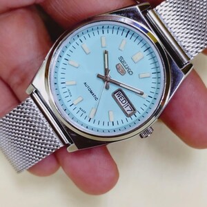K&ouml;nnte beinhalten: Eine silberfarbene Seiko 5 Automatik-Uhr mit hellblauem Zifferblatt und Mesh-Armband. Das Zifferblatt zeigt Tag und Datum, mit "MIT 17" sichtbar. Die Uhr wird von einer Hand gehalten.