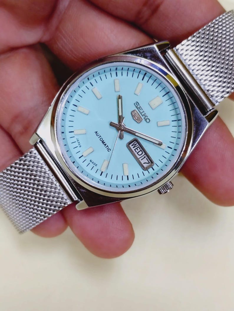 K&ouml;nnte beinhalten: Eine silberfarbene Seiko 5 Automatik-Uhr mit hellblauem Zifferblatt und Mesh-Armband. Das Zifferblatt zeigt Tag und Datum, mit "MIT 17" sichtbar. Die Uhr wird von einer Hand gehalten.