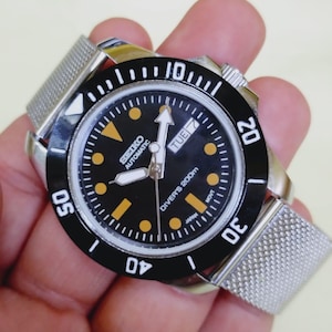 May include: A black-dial Seiko Automatic diver's watch with a silver mesh band. The watch face features luminous markers and hands, a date display, and a rotating bezel. The watch is marked "Diver's 200m" and "Japan".