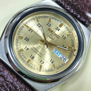 May include: Close-up of a gold-toned Seiko 5 automatic watch with a silver case and a brown patterned strap. The watch face displays the day and date, with the day reading "SAT 26". The watch has a grid pattern on the face.