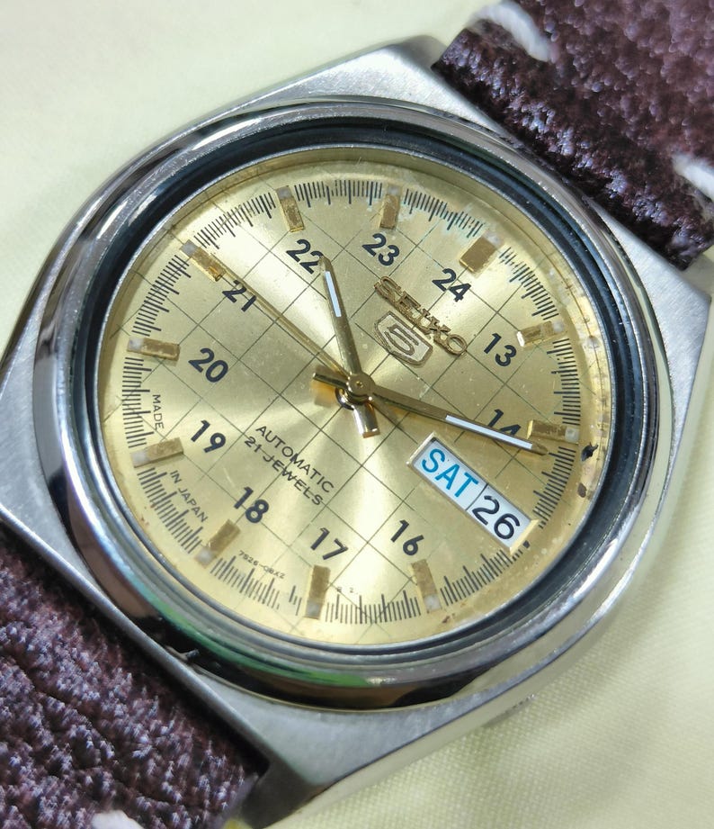 May include: Close-up of a gold-toned Seiko 5 automatic watch with a silver case and a brown patterned strap. The watch face displays the day and date, with the day reading "SAT 26". The watch has a grid pattern on the face.