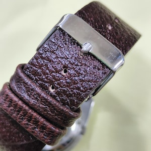May include: Close-up of a brown leather watch strap with a silver buckle. The strap has a textured surface and is secured with a metal clasp. The watch face is partially visible.